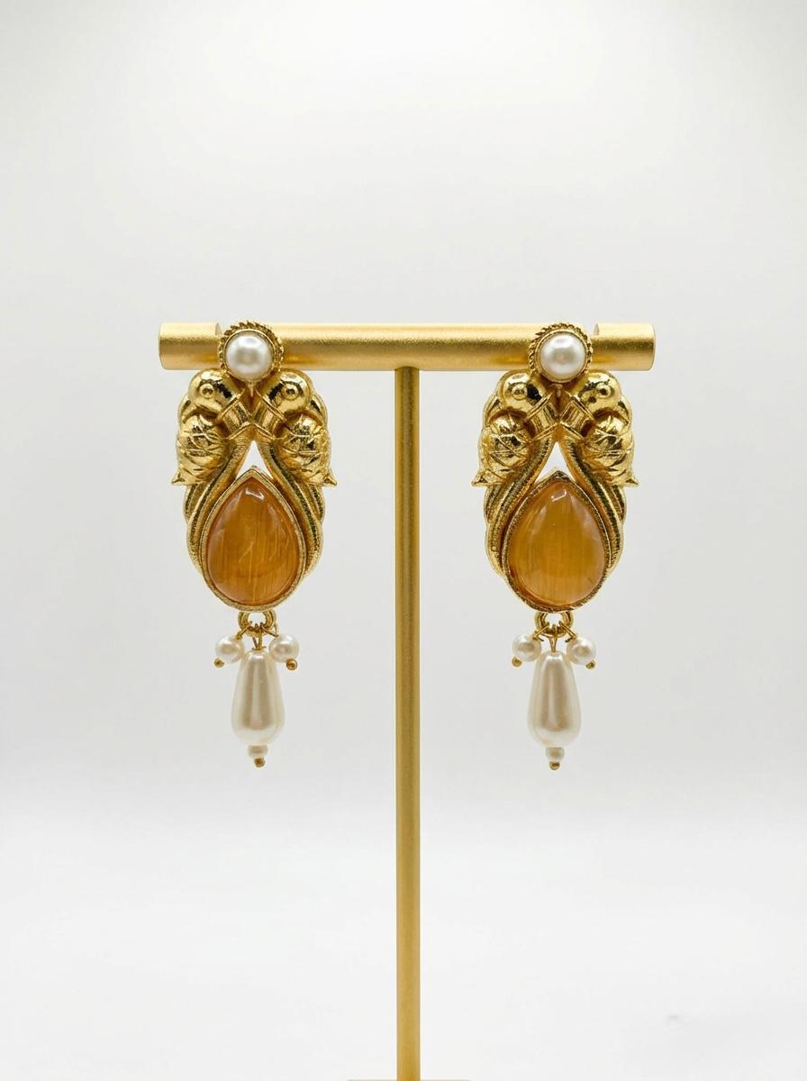 A pair of elegant all-gold jhumka drop earrings on a display stand, featuring a metallic gold cabochon finish within an ornate brass frame, complemented by traditional pearl drop detailing.