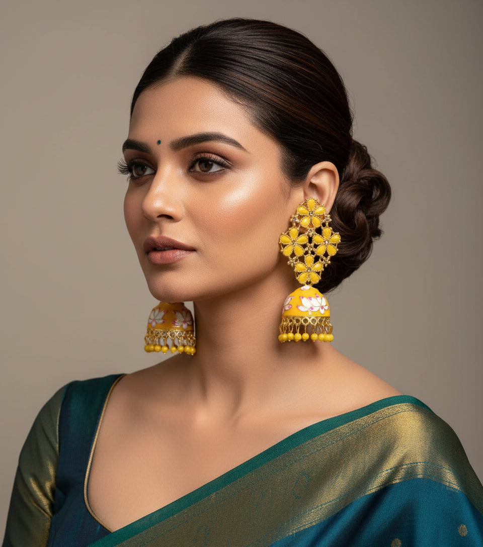 A pair of gold-plated brass jhumka earrings with yellow stones and gold meenakari work displayed on a stand.