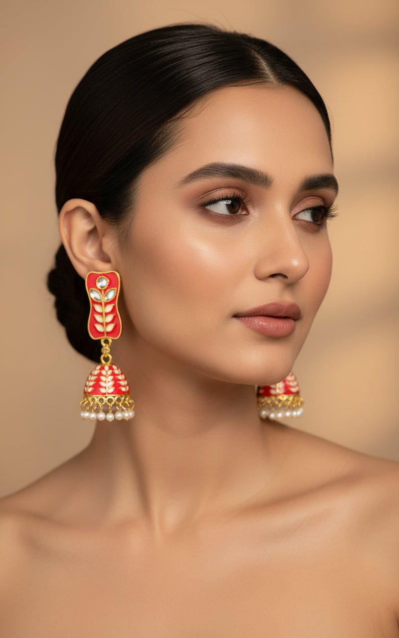 A pair of gold-plated red colour Meenakari drop Jhumka earrings displayed on a stand, featuring intricate designs and studded with stones.