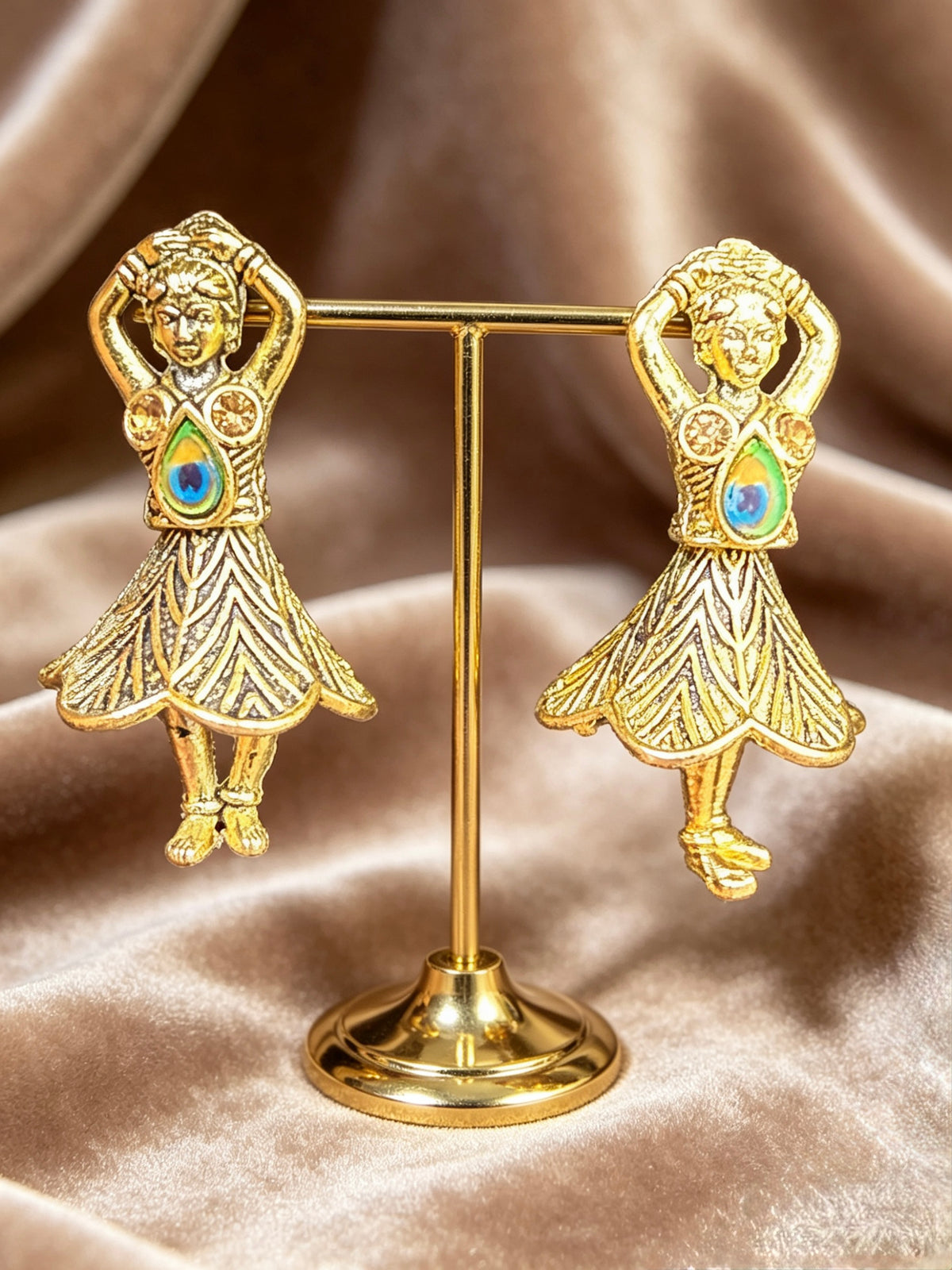 A pair of gold-toned earrings shaped like dancing girls, displayed on a small gold stand against a draped satin fabric background.