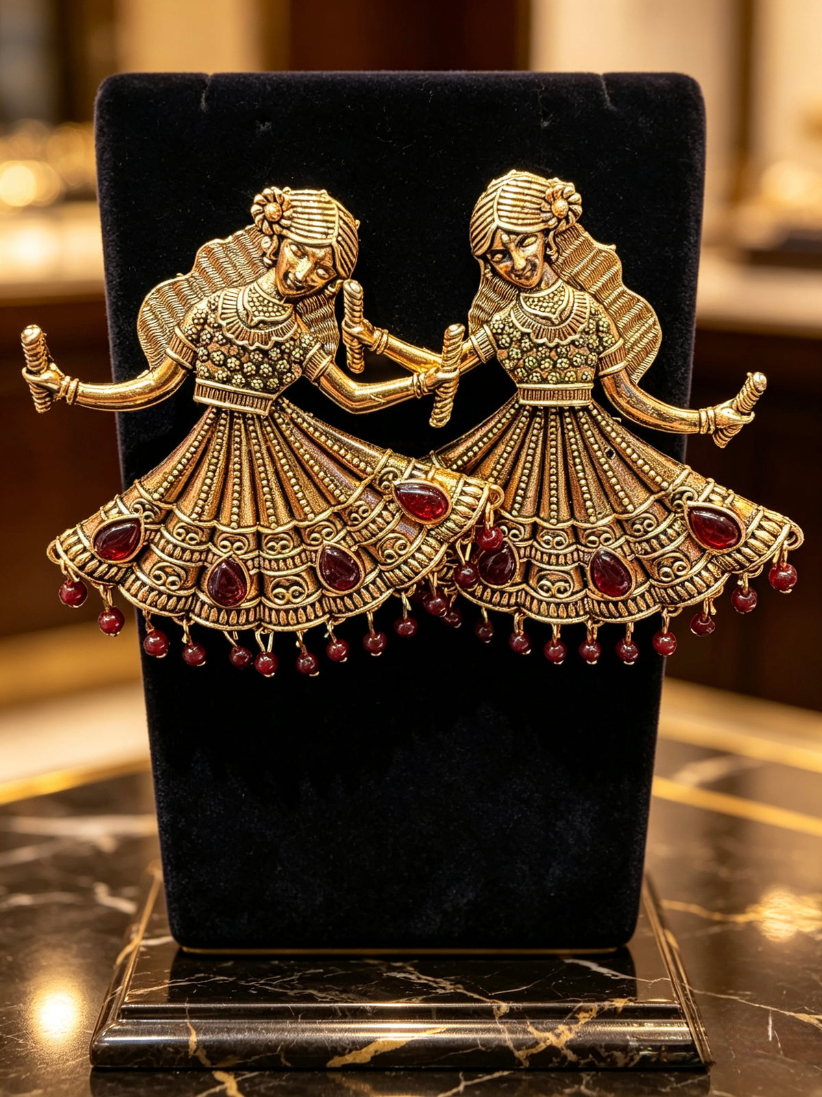 A pair of gold-toned traditional Indian bridal earrings shaped like dancing bride and groom, embellished with red teardrop gemstones and small red bead drops, displayed on a black velvet jewelry stand.