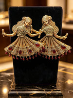 A pair of gold-toned traditional Indian bridal earrings shaped like dancing bride and groom, embellished with red teardrop gemstones and small red bead drops, displayed on a black velvet jewelry stand.