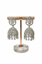 A pair of large, silver-toned bridal Jhumka earrings with high-shine crystal encrustation and hanging crystal droplets on a display stand.