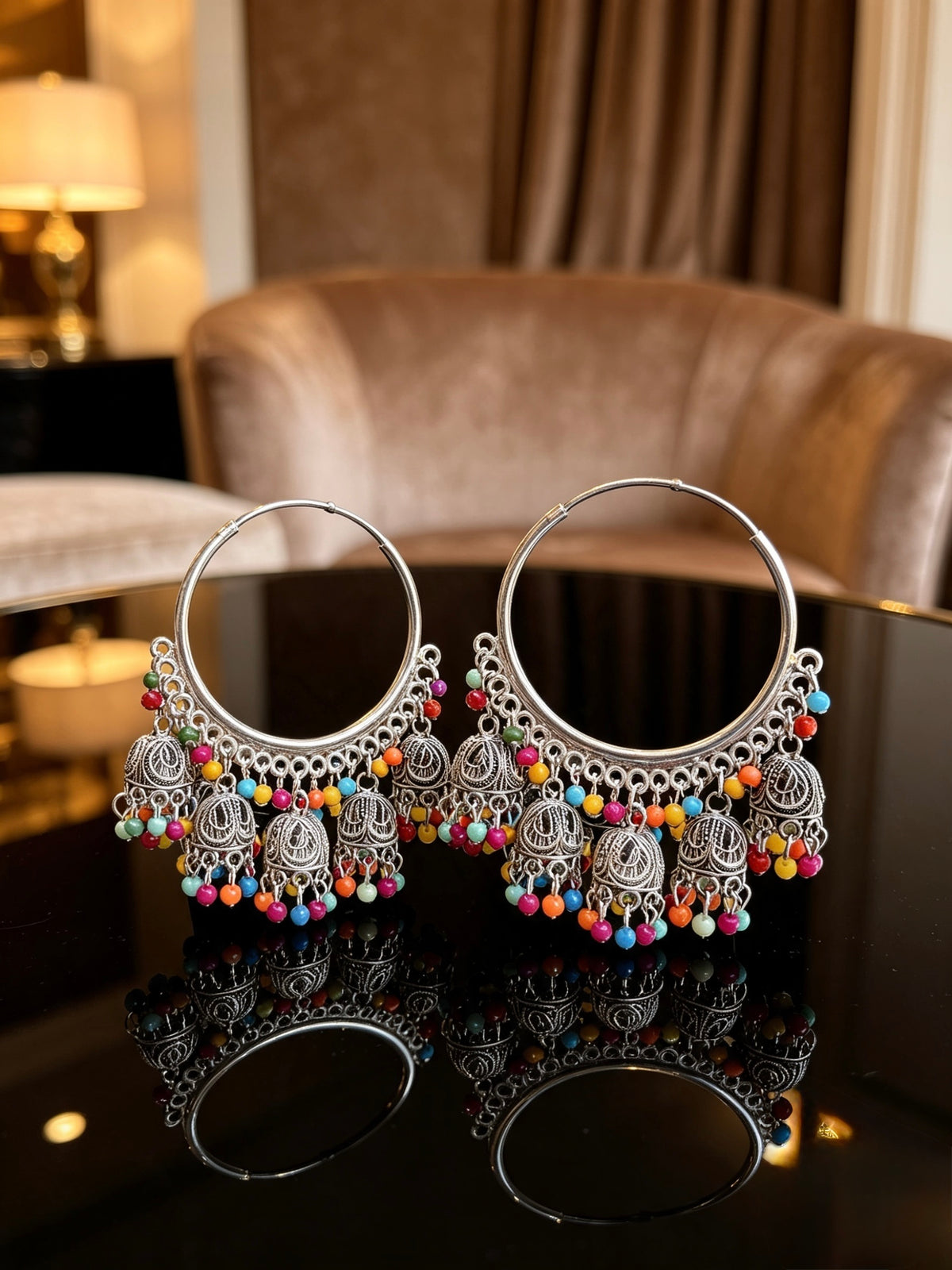 A pair of large silver-toned hoop earrings with ornate filigree dangles and colorful beaded fringe, displayed on a glossy black table with a blurred indoor lounge setting in the background.