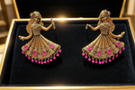 A pair of ornate Antique gold-toned earrings shaped like dancing figures, displayed in a black jewellery box. Each earring features detailed filigree, pink gemstone accents, and dangling pink bead fringe.