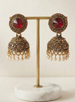 A pair of ornate Indian jhumka earrings with red circular stones and gold-toned filigree bells with pearl-like dangling beads, displayed on a gold stand with a white marble base.