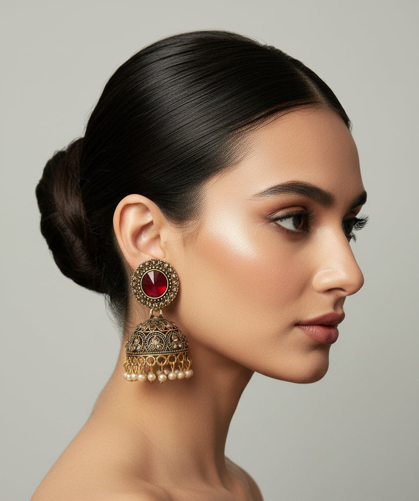 A pair of ornate gold-toned Jhumka earrings with red stones and pearl bead fringe, held on a small white card by a person's finger against a light grey background.