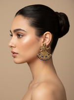 A pair of ornate gold-toned earrings shaped like dancing figures, displayed in a black jewelry box. Each earring features detailed filigree, pink gemstone accents, and dangling pink bead fringe.
