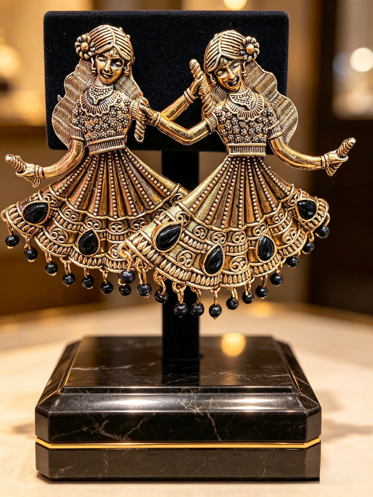 A pair of ornate gold-toned earrings shaped like dancing figures in traditional attire, embellished with black stones and hanging beads, displayed on a black stand with a black velvet backing and a black marble base.
