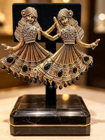 A pair of ornate gold-toned earrings shaped like dancing figures in traditional attire, embellished with black stones and hanging beads, displayed on a black stand with a black velvet backing and a black marble base.