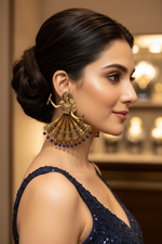 A woman in profile wearing a large ornate gold-toned traditional jhumka earring with dangling blue bead accents, photographed in a softly lit indoor setting.