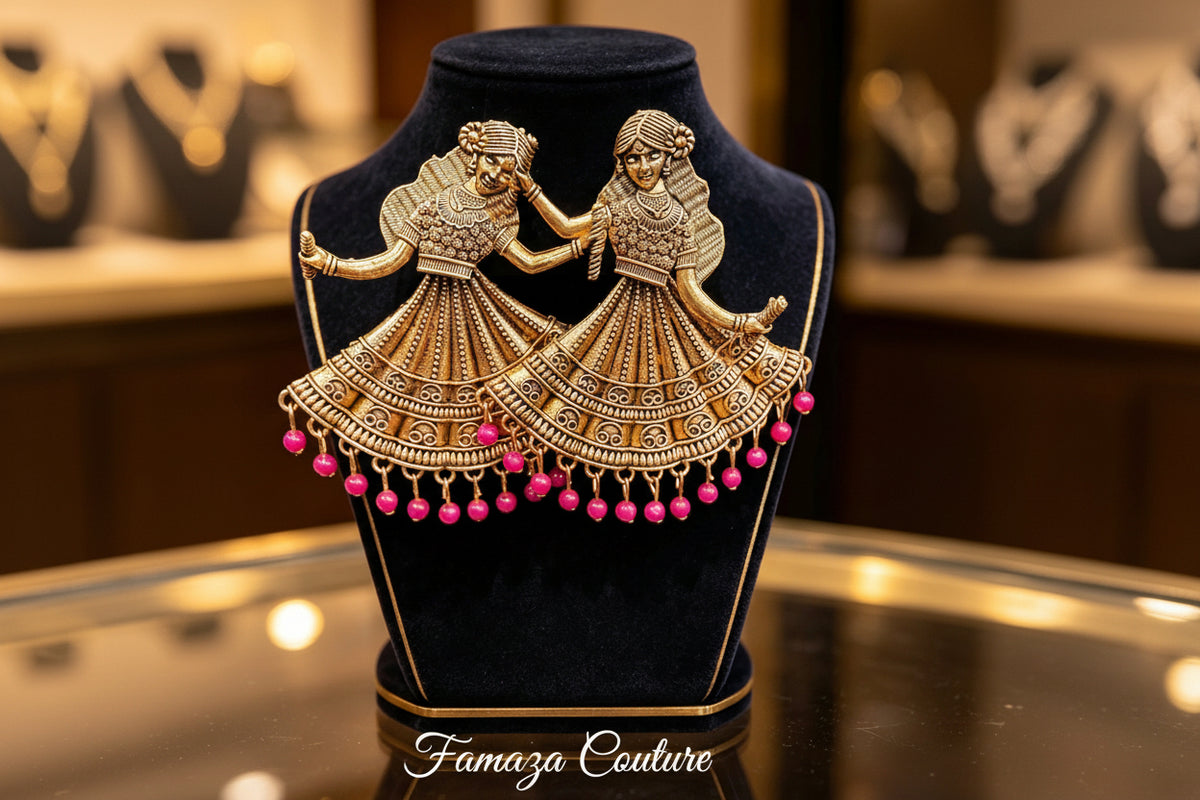 A pair of ornate gold-toned earrings shaped like dancing figures with layered skirt patterns and dangling pink bead drops, displayed on a black velvet jewellery bust.