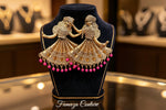 A pair of ornate gold-toned earrings shaped like dancing figures with layered skirt patterns and dangling pink bead drops, displayed on a black velvet jewellery bust.
