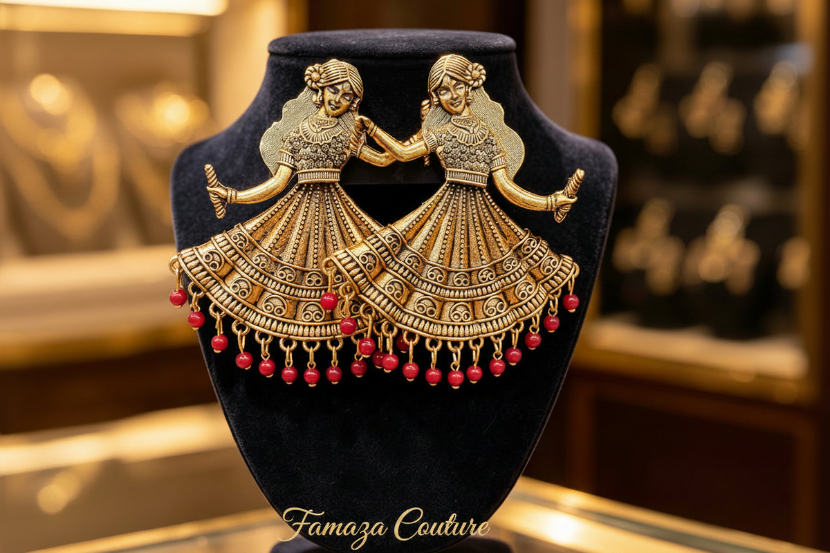 A pair of ornate gold-toned earrings shaped like two dancing figures in traditional dress, with dangling red bead chains, displayed on a black velvet jewelry bust. The background is a softly blurred warm-toned jewelry store interior.