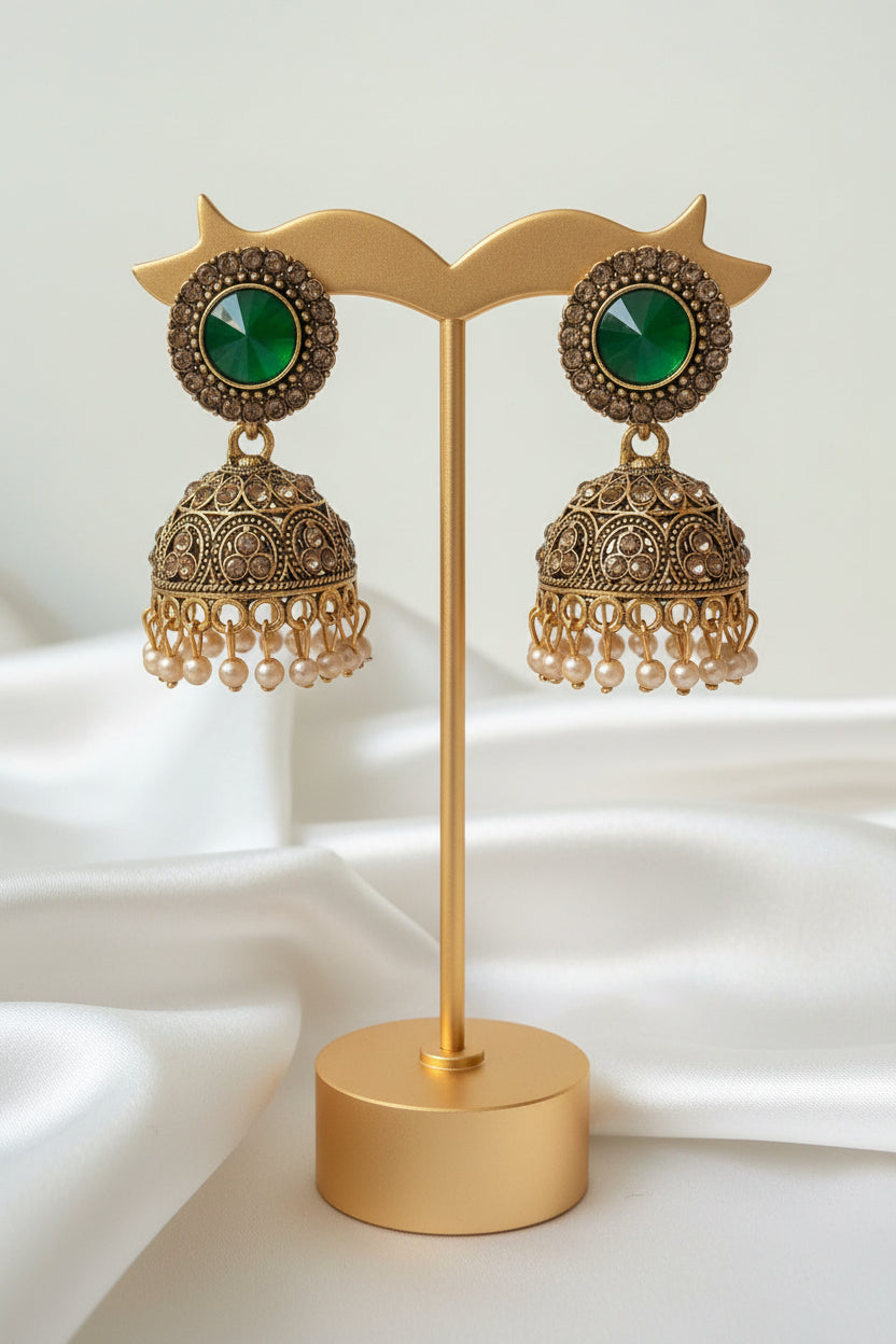 A pair of ornate gold-toned jhumka earrings with green oval stones and rhinestone borders, attached to a white jewellery display card held by a hand.