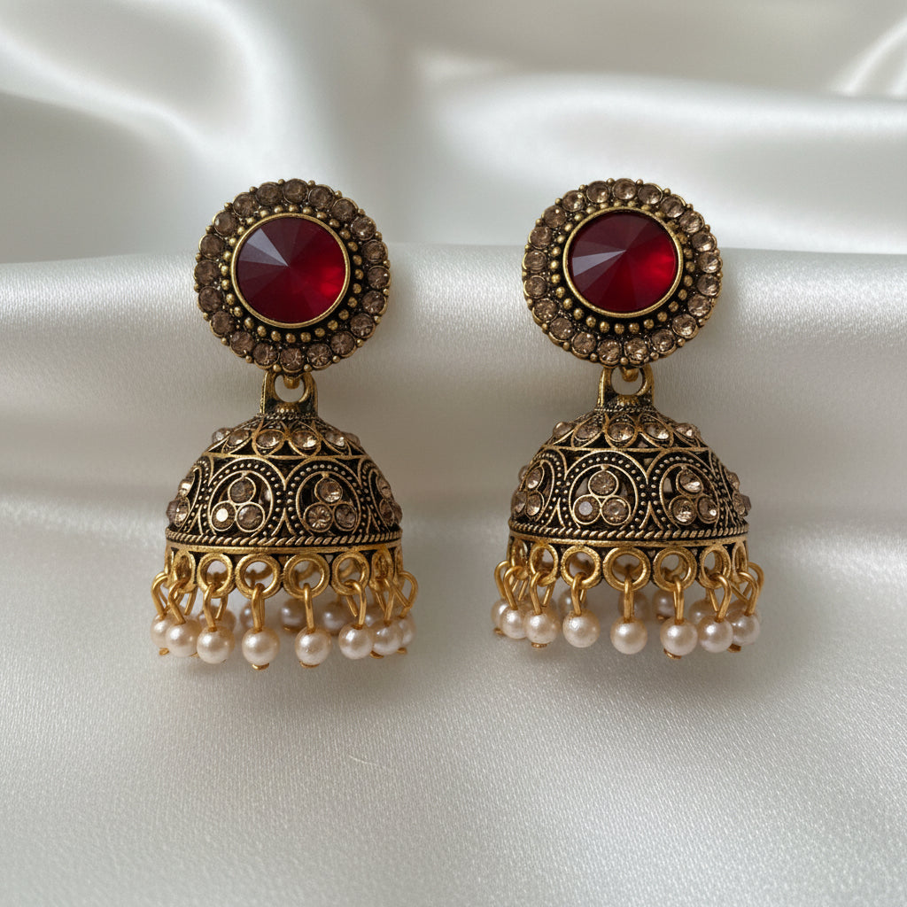 A pair of ornate gold-toned jhumka earrings with red faceted centre stones and small pearl-like drops, displayed on a white jewellery card held by a hand.
