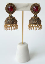 A pair of ornate gold-toned jhumka earrings with red faceted stones and clear rhinestone borders, attached to a white jewellery display card held by a hand.