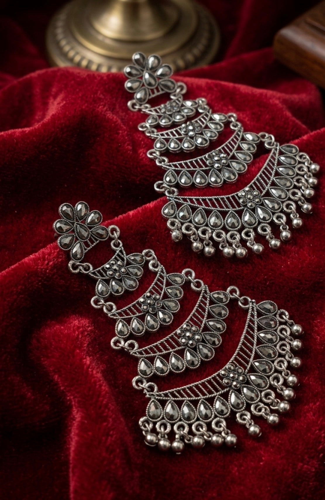 A pair of ornate silver-toned Indian jhumka earrings with intricate filigree and beadwork, displayed on a rich red velvet fabric background.