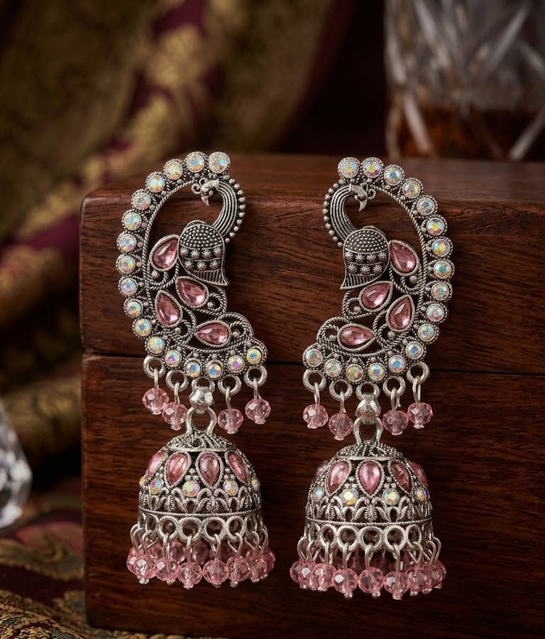 A pair of ornate silver-toned Indian jhumka earrings with pink teardrop stones and iridescent bead accents, displayed on a dark wooden box.