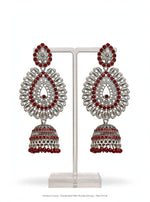 A pair of ornate silver-toned Jhumka earrings with red gemstone accents, displayed on a clear acrylic stand against a white background.