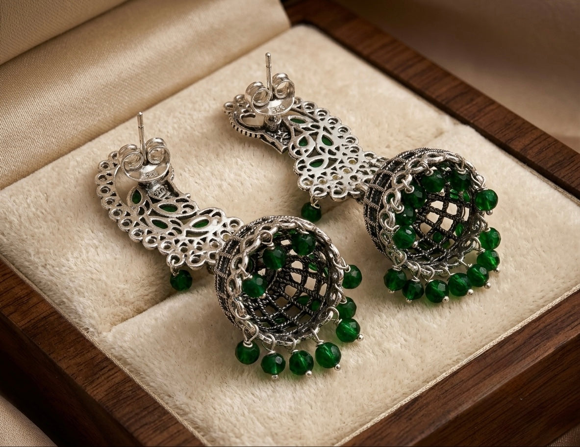 A pair of ornate silver-toned dangle earrings with intricate filigree detailing and dangling green bead accents, displayed on a beige velvet-lined jewellery box.