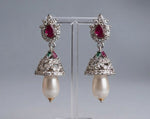 A pair of ornate silver-toned earrings with red teardrop gemstones, small green accent stones, and dangling pearl-like drops, displayed on a clear stand against a soft grey background.