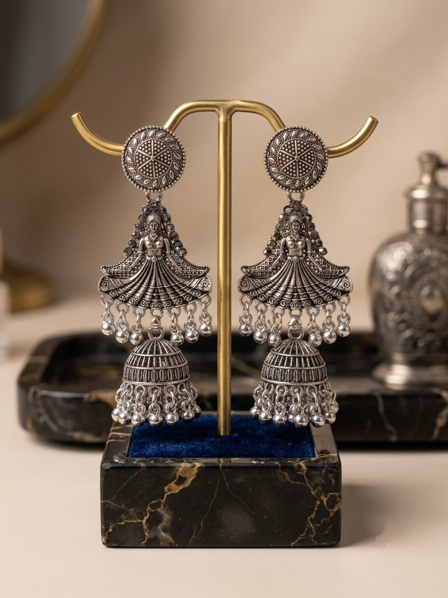 A pair of ornate silver-toned jhumka earrings displayed on a gold-tone stand with a dark marble base, set against a softly blurred warm neutral background.