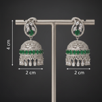 A pair of ornate silver-toned jhumka earrings with green stone accents, displayed on a silver T-stand against a dark gray background.