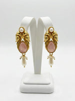  A pair of pink, vintage-style brass jhumka earrings displayed on a jewelry stand, showcasing intricate metalwork, a smooth pink cabochon center, and elegant pearl fringe detailing.