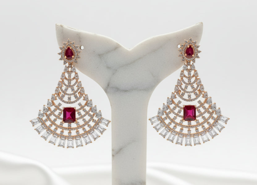 A pair of rose gold-toned chandelier earrings with red gemstone centres and clear stones, displayed on a black velvet jewellery bust inside a shop.