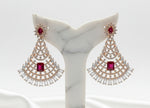 A pair of rose gold-toned chandelier earrings with red gemstone centres and clear stones, displayed on a black velvet jewellery bust inside a shop.