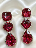 A pair of silver-tone drop earrings featuring three faceted deep red gemstones in a cascading design, displayed on a draped white satin fabric background.