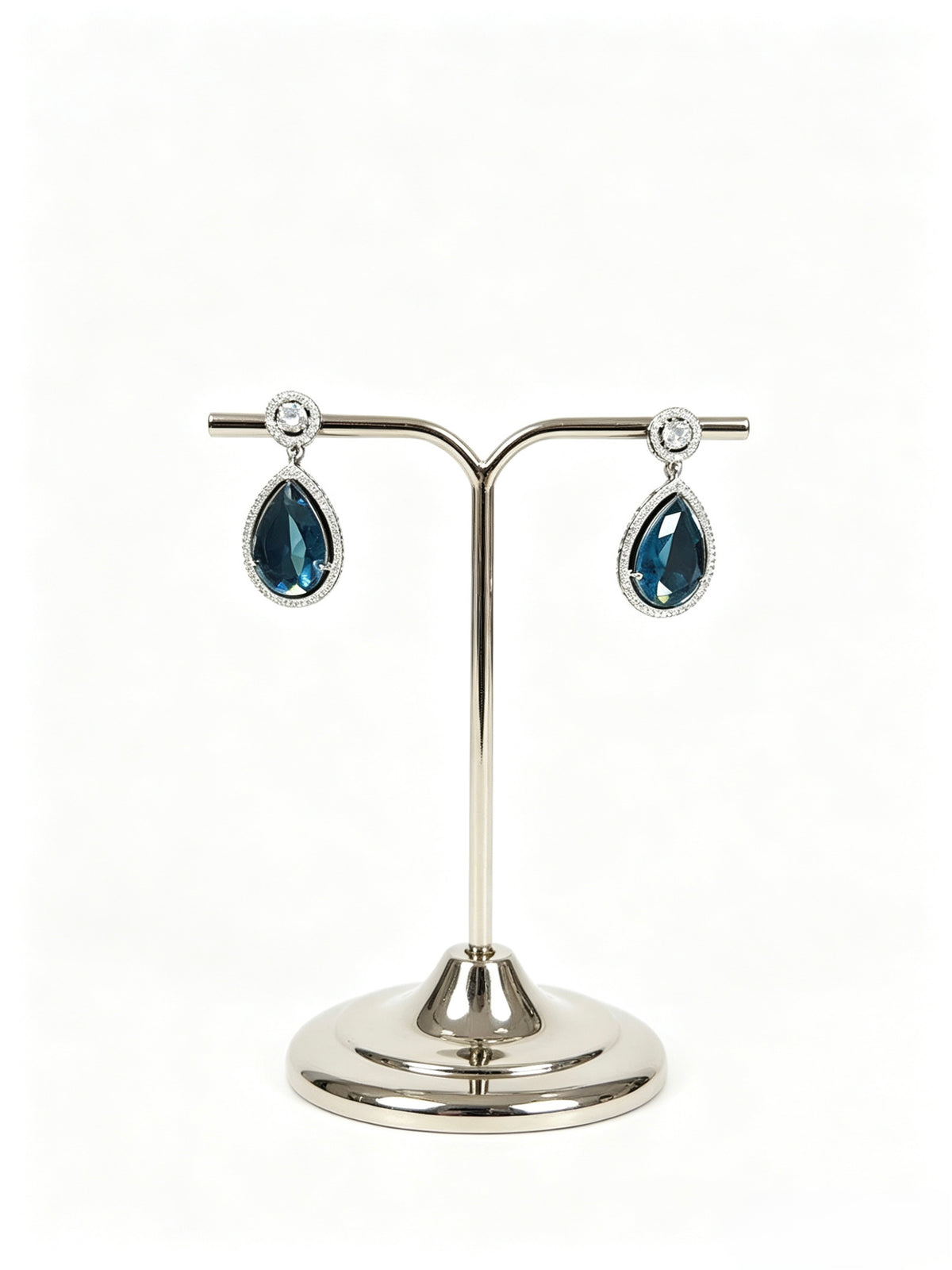 A pair of silver-tone teardrop dangle earrings with dark blue faceted gemstones and small clear stones at the top, displayed on a polished silver jewellery stand against a white background.