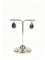 A pair of silver-tone teardrop dangle earrings with dark blue faceted gemstones and small clear stones at the top, displayed on a polished silver jewellery stand against a white background.