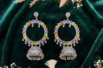 A pair of silver-toned Indian jhumka earrings with colorful enamel accents in red, blue, green, and yellow, displayed on a dark green velvet fabric background with dried floral decorations.