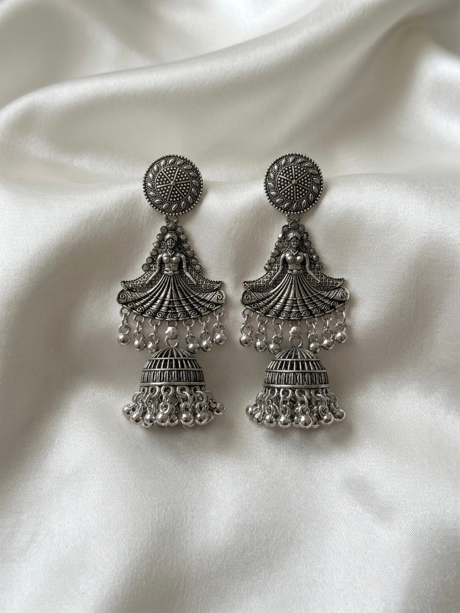 A pair of silver-toned Indian jhumka earrings with intricate filigree work and dangling bead chains, displayed on a draped white satin fabric background.