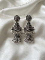 A pair of silver-toned Indian jhumka earrings with intricate filigree work and dangling bead chains, displayed on a draped white satin fabric background.