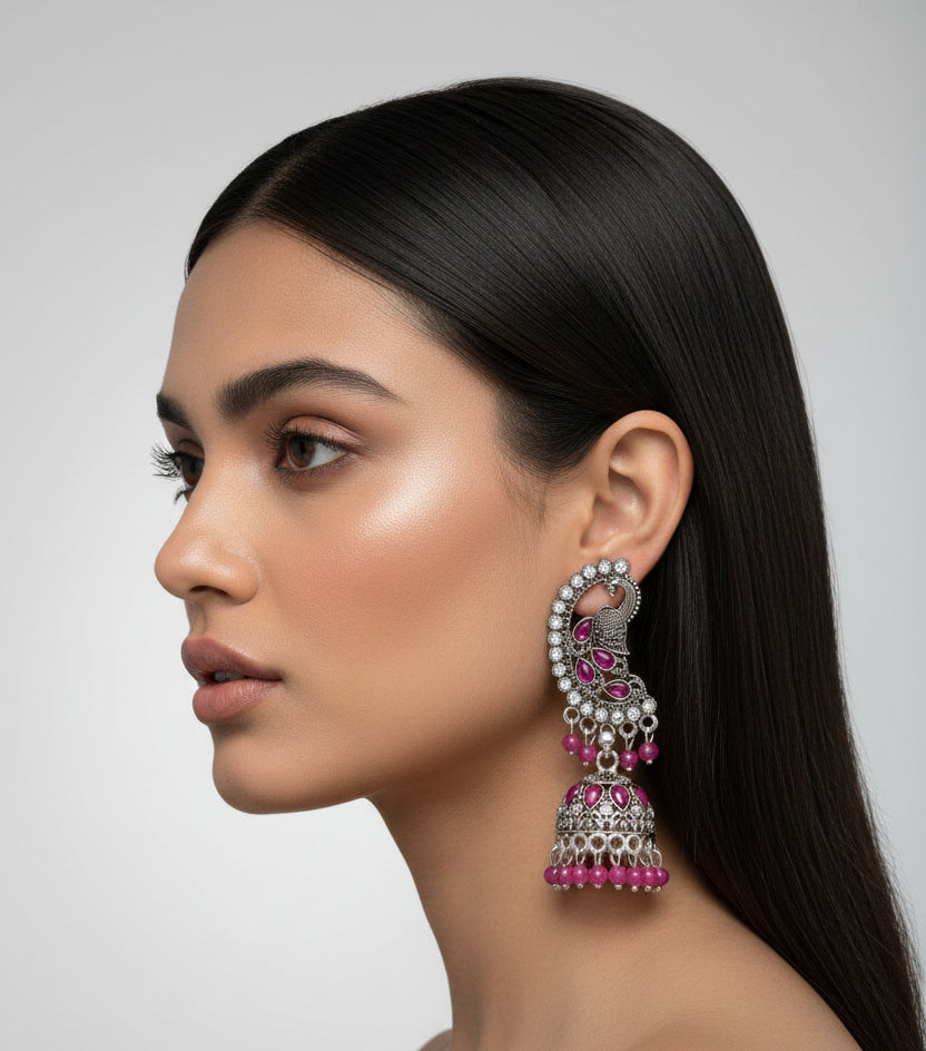 A pair of silver-toned Indian jhumka earrings with pink teardrop and round crystals, attached to a blue velvet display card held by a hand against a dark textured background.