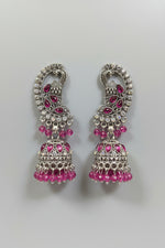 A pair of silver-toned Indian jhumka earrings with pink teardrop and round crystals, attached to a blue velvet display card held by a hand against a dark textured background