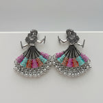 A pair of silver-toned dholak dancer earrings with multi-coloured beaded skirts, displayed on a white marble surface.