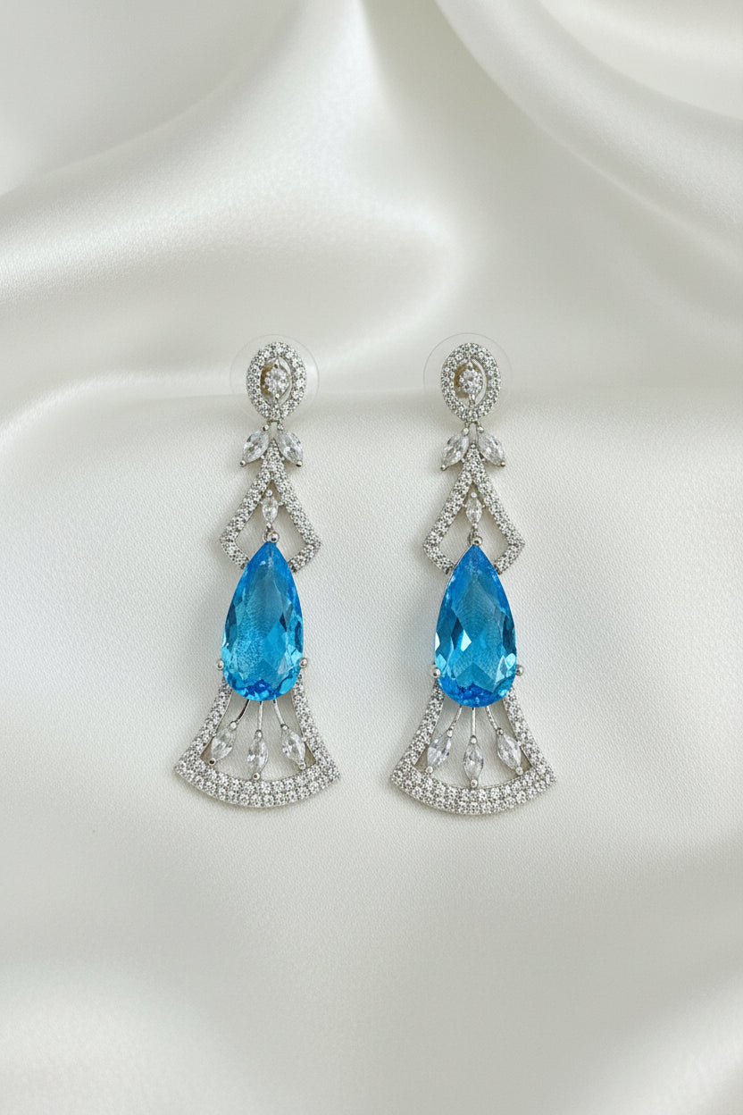 A pair of silver-toned drop earrings with a central faceted blue marquise-cut gemstone, surrounded by smaller clear stones in an Art Deco-inspired fan and leaf design, displayed on a black textured background.