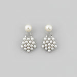 A pair of silver-toned drop earrings with a single round white pearl and a small cluster of tiny silver pearls, displayed on a light wood floor.