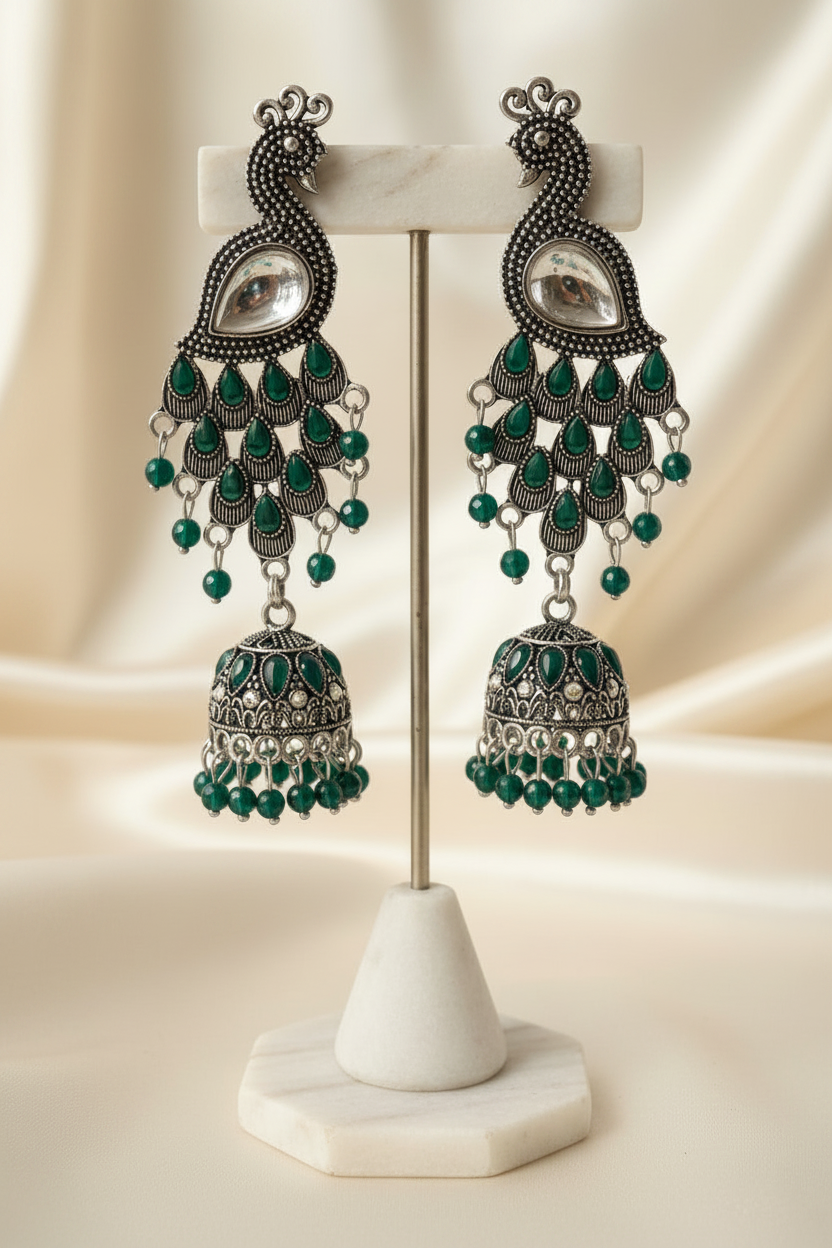 A pair of ornate silver-toned dangle earrings with green teardrop and bead details, displayed on a white stand against a beige satin background.