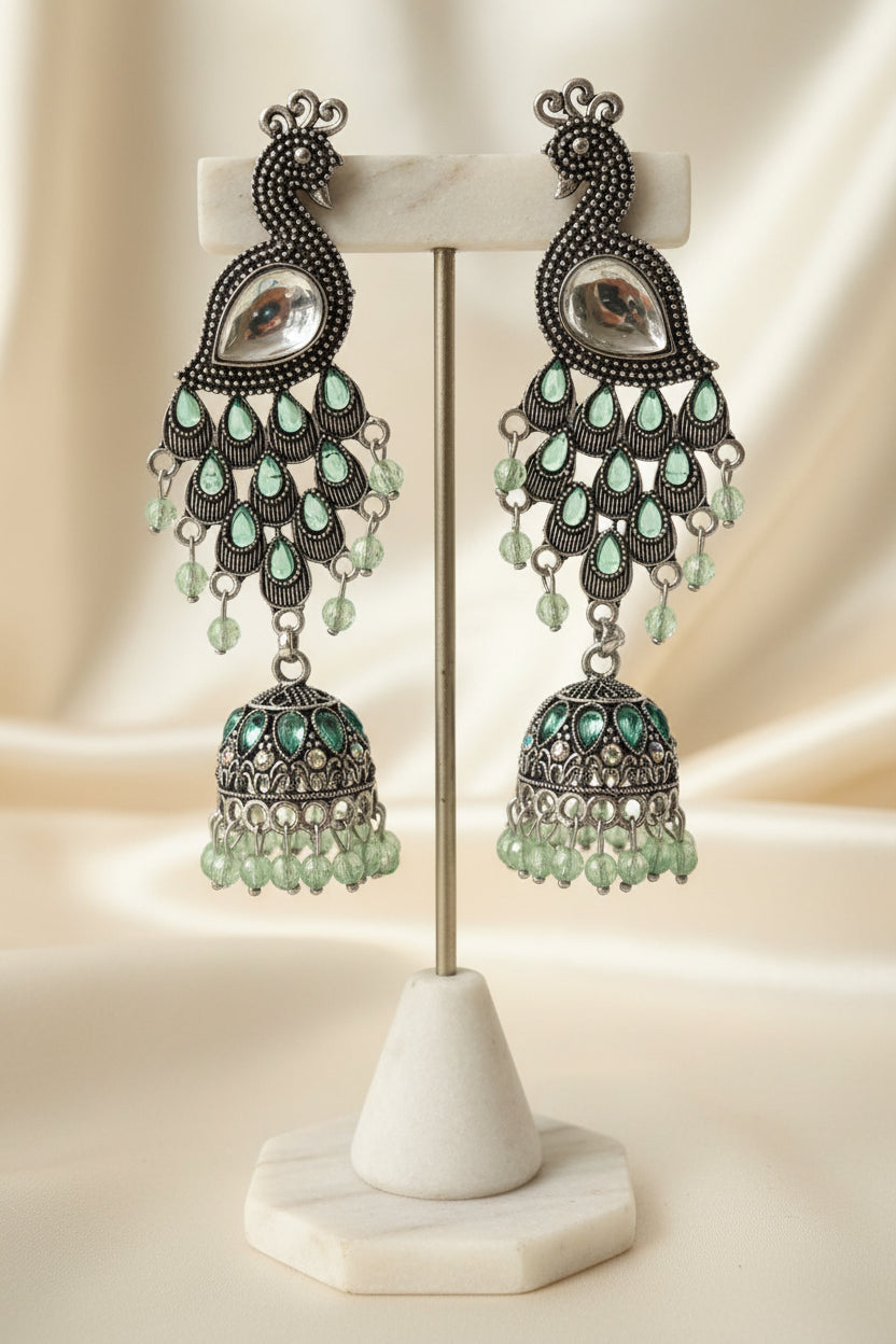 A pair of silver-toned peacock jhumka earrings with green teardrop rhinestones and dangling green beads, attached to a blue display card and held by a hand against a plain light background.