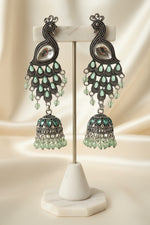 A pair of silver-toned peacock jhumka earrings with green teardrop rhinestones and dangling green beads, attached to a blue display card and held by a hand against a plain light background.