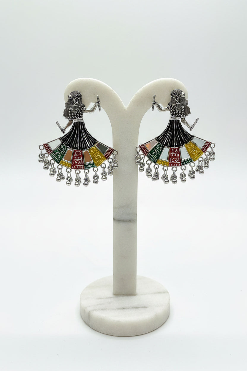 A pair of silver-toned statement earrings shaped like dancing women with colorful geometric dress details and dangling silver beads, held in a hand against a light marble background.