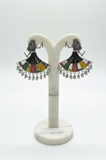 A pair of silver-toned statement earrings shaped like dancing women with colorful geometric dress details and dangling silver beads, held in a hand against a light marble background.
