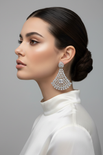 A woman in profile wearing a large, intricate silver-toned teardrop chandelier earring with clear gemstones, paired with a white high-neck top against a neutral gray background.