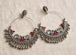 A pair of silver-toned traditional hoop earrings with intricate filigree, bird motifs, and small red stone accents, displayed on a textured beige fabric background.