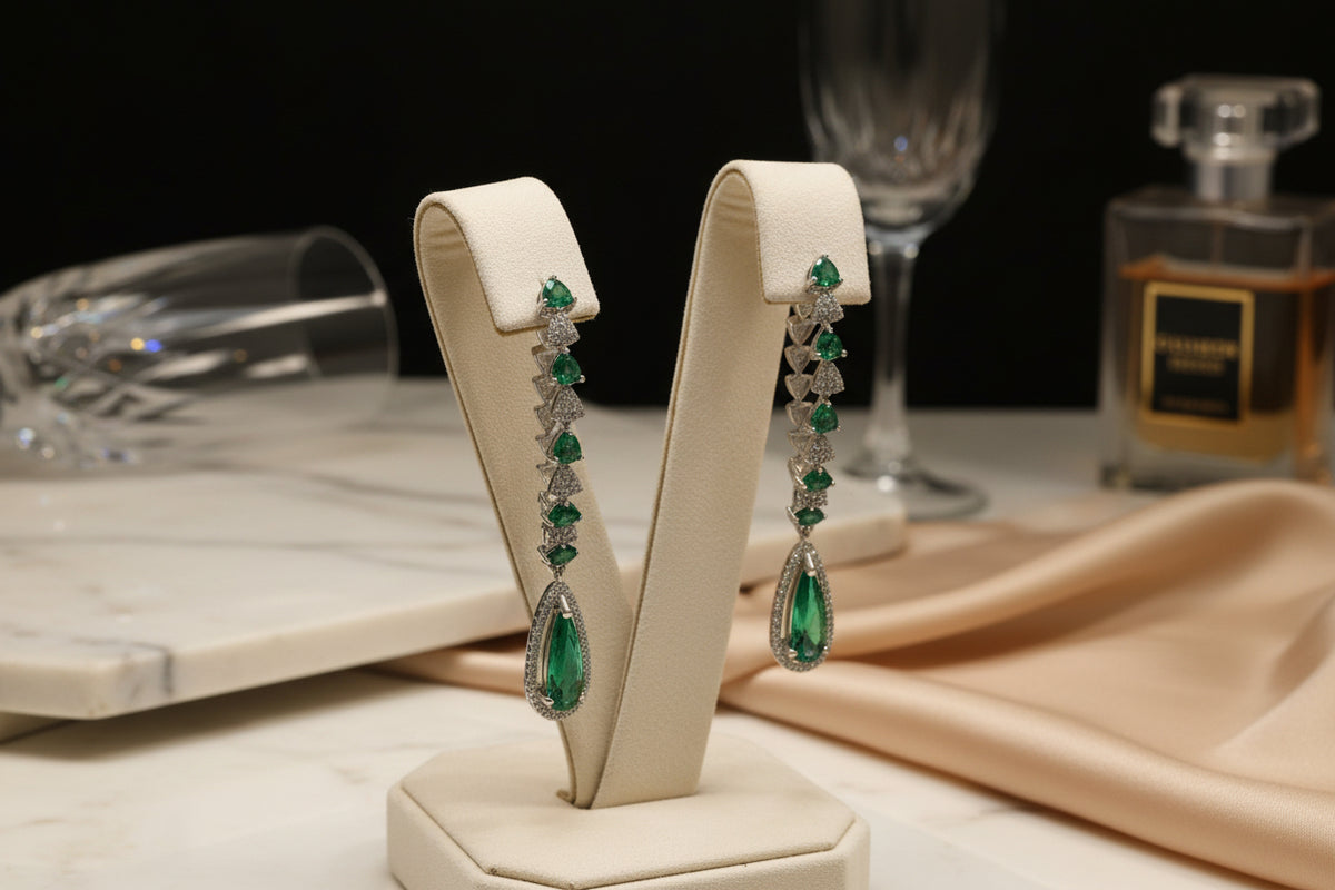 A pair of silver finish American diamond Jhumka bali earrings with green and clear stones on a stand.